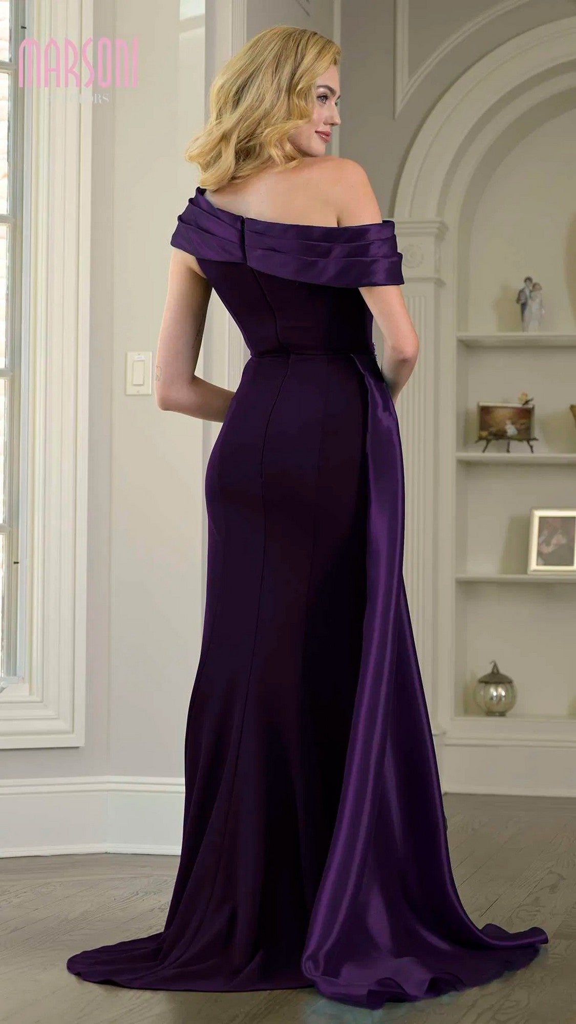 A woman stands indoors facing away, showcasing the elegant back and train of the Marsoni M346 long formal dress, an off-the-shoulder, floor-length stretch crepe gown with a fitted sheath silhouette. Shelves and decor fill the background.